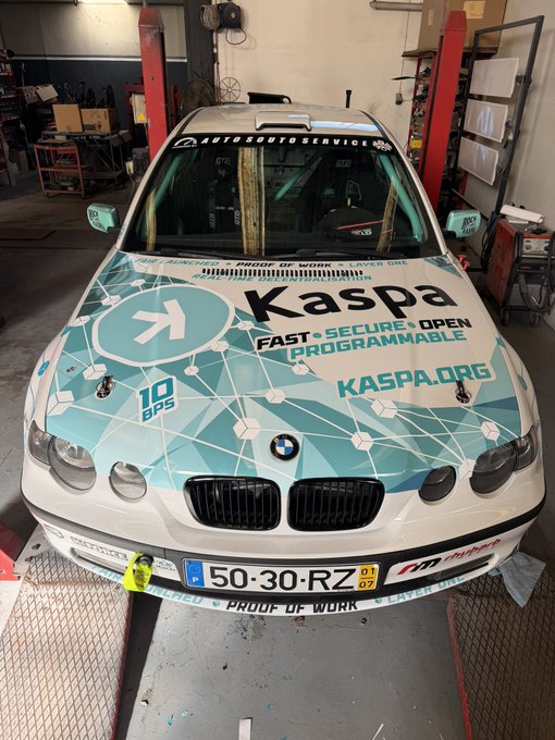 A BMW race car in a garage wrapped in teal and white Kaspa branding with 'KASPA.ORG', 'FAST SECURE OPEN PROGRAMMABLE', and '10 BPS' visible on the hood.