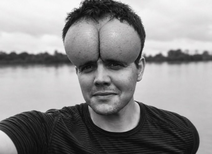 A black and white selfie of a man with an unusually large, elongated forehead/forehead protrusion, standing outdoors near a body of water.