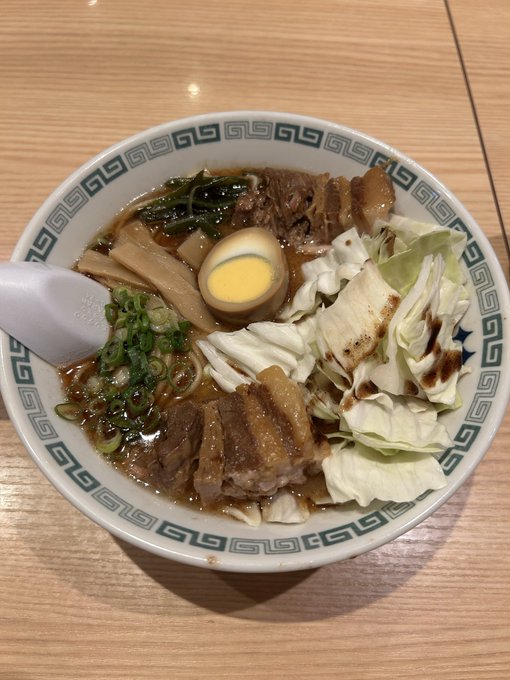 A bowl of ramen with a soft-boiled egg, beef, cabbage, bamboo shoots, and green onions, served in a traditional bowl with a Greek key pattern on the rim.
