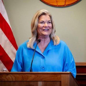 Senator Cynthia Lummis, a blonde woman in a blue blouse, stands at a podium delivering remarks, with an American flag visible behind her. The image accompanies breaking news about her urging Democrats to pass the CLARITY Act to establish federal protections, disclosure rules, and delisting requirements for the digital asset market.