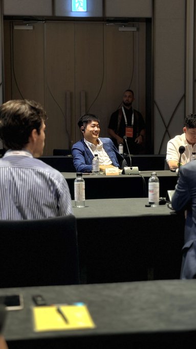 A scene from what appears to be a business meeting or panel discussion, with a smiling man in a blue blazer sitting at a table with water bottles and a microphone, while another man in a striped shirt sits in the foreground.