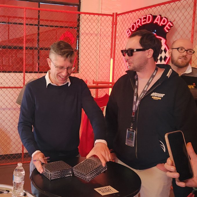 Two men wearing lanyards stand in front of a red chain-link fence with a 'BORED APE' sign; one man in a black jacket holds sunglasses while the other in a blue sweater smiles, with what appears to be NFT cards on the table before them.