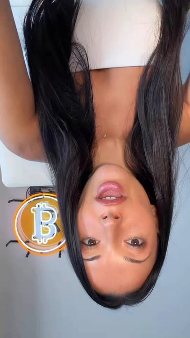 An upside-down selfie of a woman with long dark hair wearing a white top, with a Bitcoin logo sticker visible behind her.