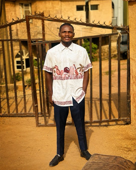 A young man stands in front of a metal gate wearing a white short-sleeved shirt with red floral embroidery and dark pants.