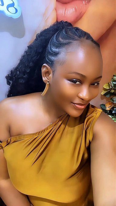 A woman with curly hair styled in a high ponytail wears a mustard yellow off-shoulder top and gold hoop earrings, posing for a selfie in front of a poster featuring pink lips.