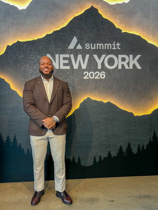 A man in a brown blazer and cream pants posing in front of a 'Summit NEW YORK 2026' backdrop featuring a mountain graphic.