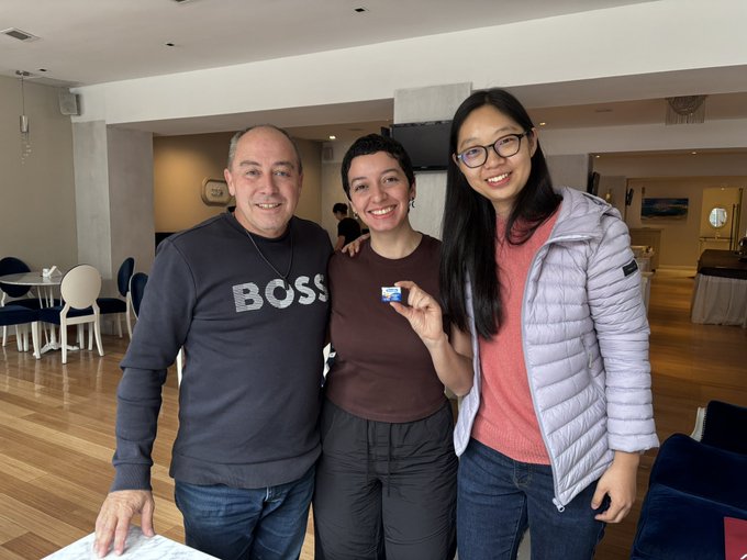 A photo of three people smiling together indoors; a man wearing a navy "BOSS" sweater on the left, a woman with short hair in the center, and a woman with glasses and a grey jacket on the right.