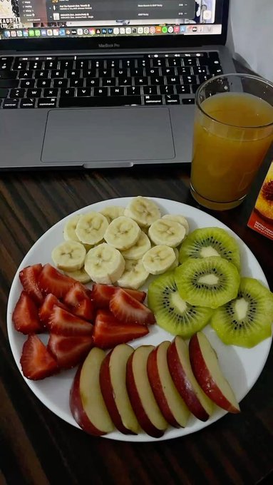 A vibrant weekend breakfast of sliced strawberries, bananas, kiwis, and apples arranged on a white plate sits beside a glass of orange juice and a MacBook Pro, perfectly capturing a relaxed and healthy start to the weekend.