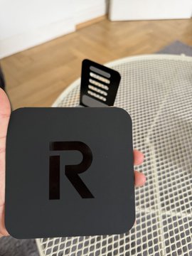 A hand holding a black Roku streaming device with the letter "R" logo, positioned over a glass patio table.