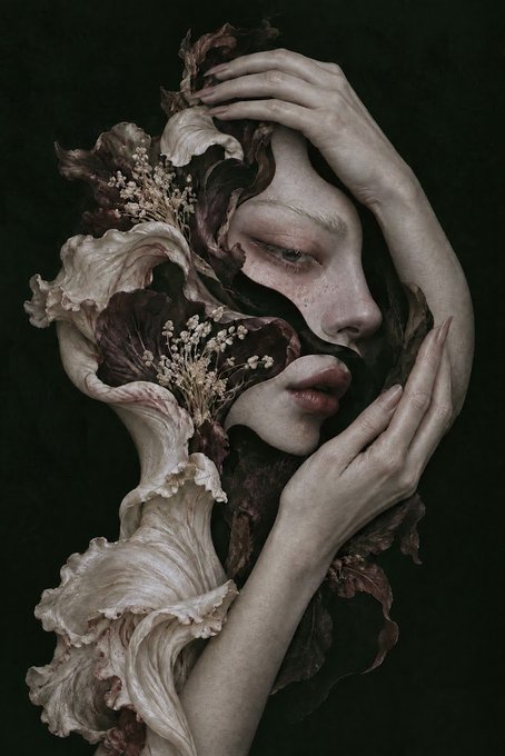 A dark, artistic portrait shows a woman with closed eyes holding her face, adorned with dried flowers, baby's breath, and dark leaves, creating a melancholic, botanical aesthetic.