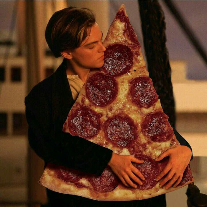 A man with brown hair in a black suit tenderly holds a large triangular slice of pepperoni pizza against his chest with his eyes closed.