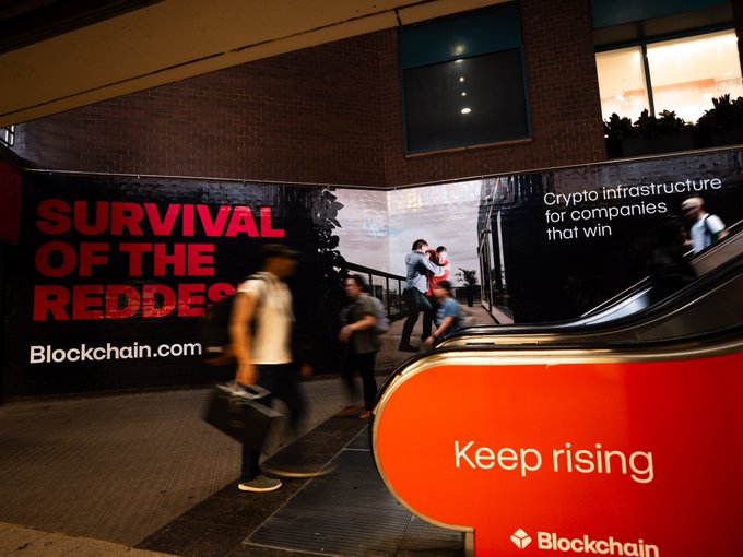 An advertisement for Blockchain.com ("Survival of the Reddest — Crypto infrastructure for companies that win — Keep rising") is displayed in a New York City subway station where commuters are walking past the escalators. The bold branding and tagline tie into the "hey NYC" post, showcasing Blockchain.com's presence in the heart of the city.