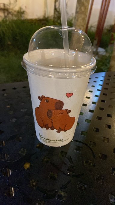 A plastic cup with a dome lid and straw, featuring a cartoon illustration of two capybaras and a heart, with the text 'kaspibara kafé' printed on it, sitting on a black metal table.
