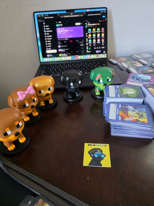A workspace featuring a laptop with a Discord window open, accompanied by several collectible figurines, stacks of trading cards, and a yellow NFT card displayed on the wooden desk.