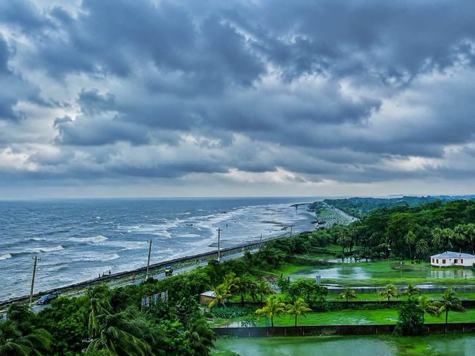 An aerial landscape view of a stormy coastline with dark clouds, ocean waves, a coastal road, palm trees, and flooded green fields.
