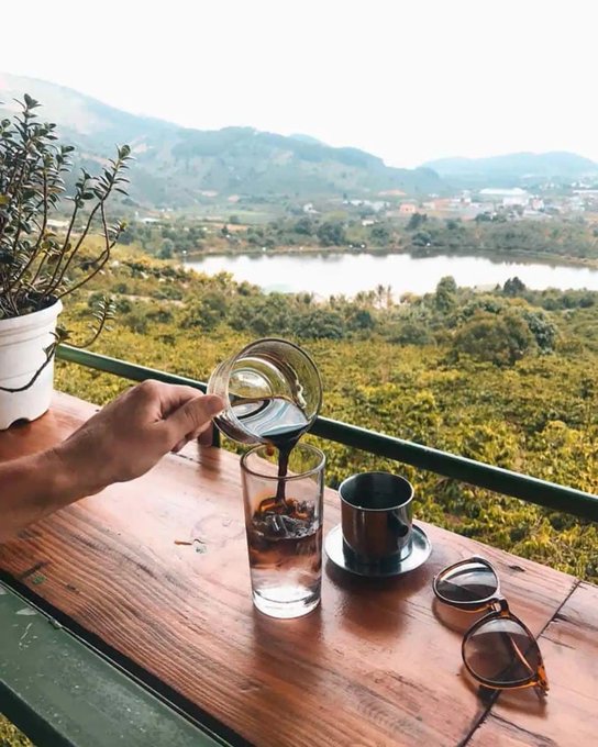A hand pours dark coffee from a glass carafe into a glass on a wooden balcony table, with a scenic view of a river, forested hills, and a village in the background.