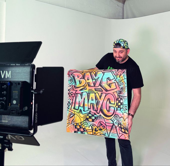 A man wearing a colorful patterned cap and a black t-shirt holds up a vibrant graffiti-style artwork featuring the text 'DAZE MA' in a studio setting.