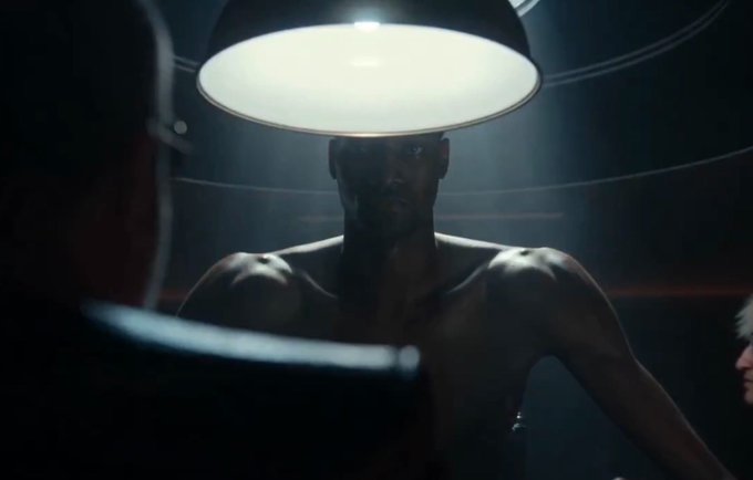 The image features a shirtless man with black hair, standing in front of a large light fixture that appears to be turned off or dimmed. There are two other people present in the scene, one located at the top left corner and another person towards the bottom right corner. The background is dark, which contrasts with the bright light from the lamp. The image also contains text that reads "solana".