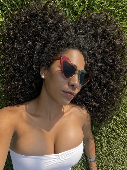 A selfie of a woman with voluminous curly hair lying on green grass, wearing heart-shaped rainbow sunglasses, pearl earrings, and a white top.