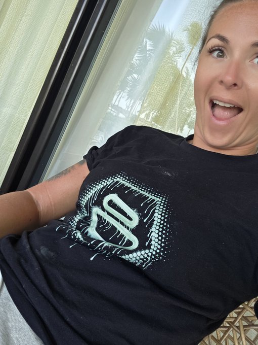 A woman in a black graphic t-shirt smiles excitedly while taking a selfie, with palm trees visible through a window behind her, fitting the "$ZBCN in the Dominican Republic" post.