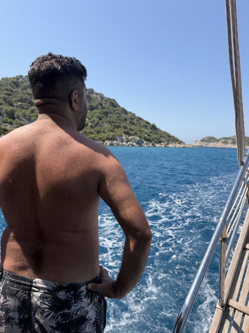 A shirtless man with short dark hair and a beard stands on the back of a boat, looking out over blue ocean water toward a green, hilly coastline under a clear sky.