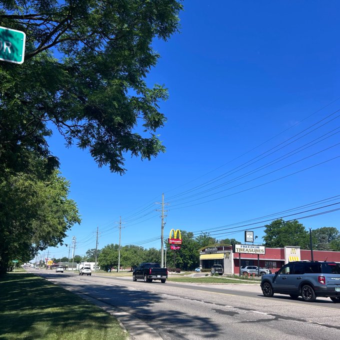 The image shows a street scene with cars driving down the road, including one car that appears to be moving away from the camera. The sky above has a blue color, suggesting a clear day. There are also trees visible along the street, adding greenery to the urban setting. A green sign is present in the scene, possibly indicating directions or information about nearby businesses and attractions.