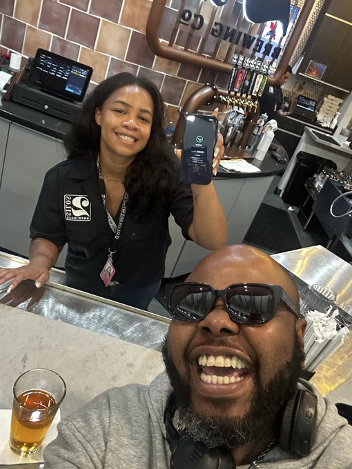 A selfie of a man at a bar taking a photo with a smiling woman behind the counter who is holding up her phone displaying a green WhatsApp-style notification screen.