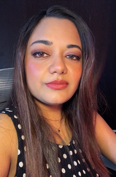 Selfie-style portrait of a woman with long brown hair, wearing makeup and a black polka-dot top, sitting in front of a dark background.