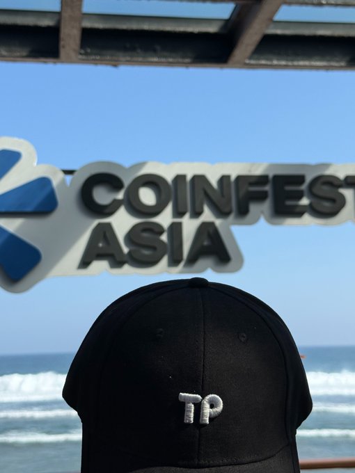 A black cap with "TP" embroidered in white sits in the foreground with the "COINFEST ASIA" sign and ocean visible in the background.