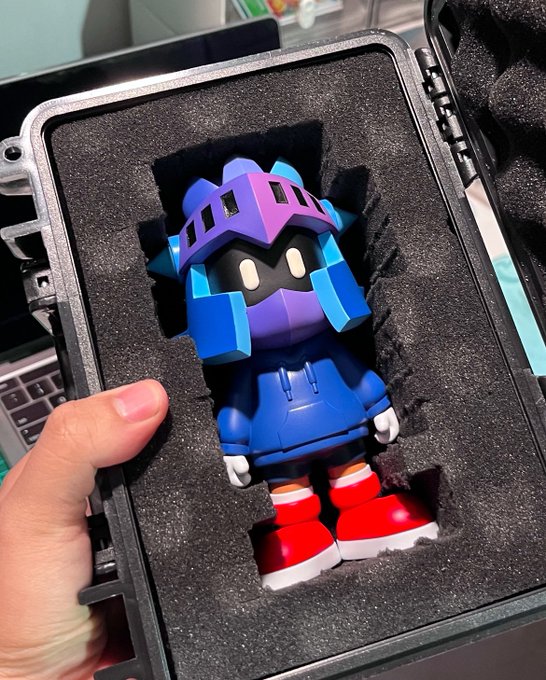 A hand holds open a black hard case displaying a blue and purple robot figurine with red and white sneakers nestled in foam padding.
