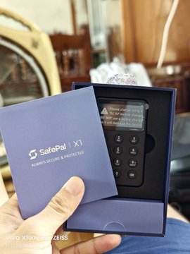 A hand holding a SafePal X1 hardware wallet in its dark blue packaging, with the device screen displaying 'Double-click to enter the set up mode'.