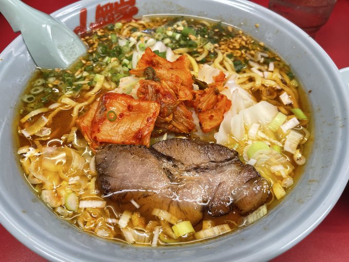 A close-up photo of a bowl of ramen featuring chashu (braised pork belly), kimchi, scallions, bamboo shoots, and noodles in a brown broth, with a white spoon resting in the bowl on a red surface.