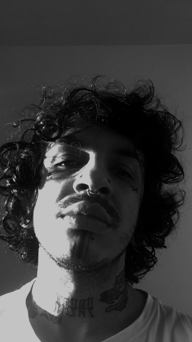 A black-and-white photograph of a young man with curly hair, a nose ring, facial tattoos, and visible neck tattoos, looking at the camera.