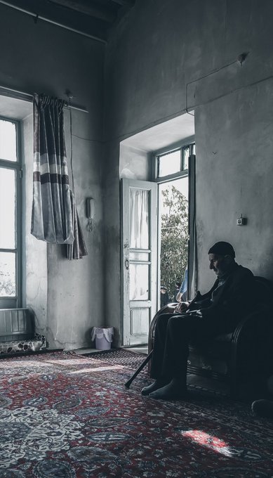 An elderly man sits silhouetted by a doorway in a sparsely furnished, high-ceilinged room, bathed in soft, muted light that emphasizes his solitude. The image evokes "The Last Sun," a contemplative NFT piece priced at 1 tez on objkt.com, capturing the quiet stillness of a lone figure in a nostalgic, almost cinematic setting.