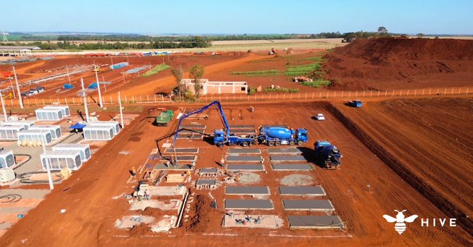 An aerial view shows a large-scale construction site with a concrete pump truck and other heavy equipment working on the foundations of industrial buildings amid expansive reddish-brown earth, with several white container-like structures on the left and a "HIVE" watermark with a bee logo in the bottom right corner. The image relates to the accompanying post by depicting the physical infrastructure development of HIVE Digital Technologies, highlighting the company's ongoing expansion projects that may have attracted the interest of investor Leopold Aschenbrenner.
