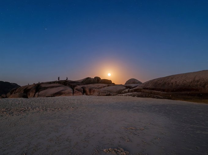 Two small figures stand atop a rocky outcrop beneath a moonlit sky, sharing the same luminous moon that inspires the post's reflection on connection across distance. The serene coastal scene, with its glowing moon rising over weathered boulders, visually echoes the message's theme of longing and shared wonder between separated souls.