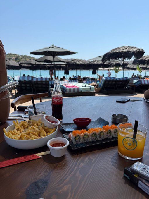 A beachfront dining scene featuring a wooden table set with french fries, Coca-Cola, sushi rolls, sauces, and an orange juice drink, with a sandy beach, umbrellas, and ocean visible in the background.