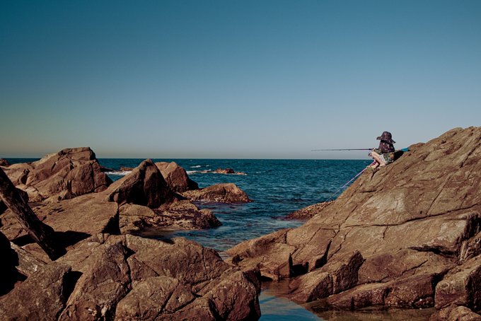 A person wearing a witch hat sits fishing on large rocky cliffs overlooking a deep blue ocean under a clear sky.