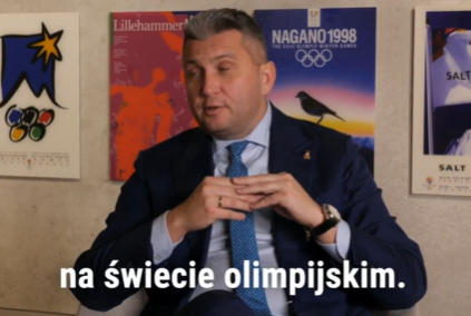 A man in a dark suit and blue tie sits in front of Olympic posters, with Polish subtitles reading 'na świecie olimpijskim.'