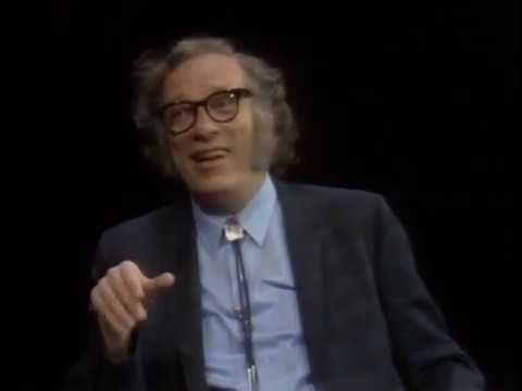 An older man with curly hair and round glasses, wearing a suit and bolo tie, speaks while gesturing with his hand.