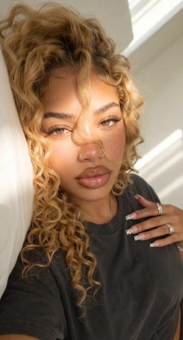 A close-up selfie of a woman with curly blonde hair, a nose ring, freckles, and manicured nails wearing a dark shirt, with sunlight casting shadows across her face.