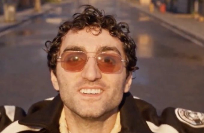 A man with curly dark hair and orange-tinted sunglasses wearing a black and white jacket, smiling on a street.
