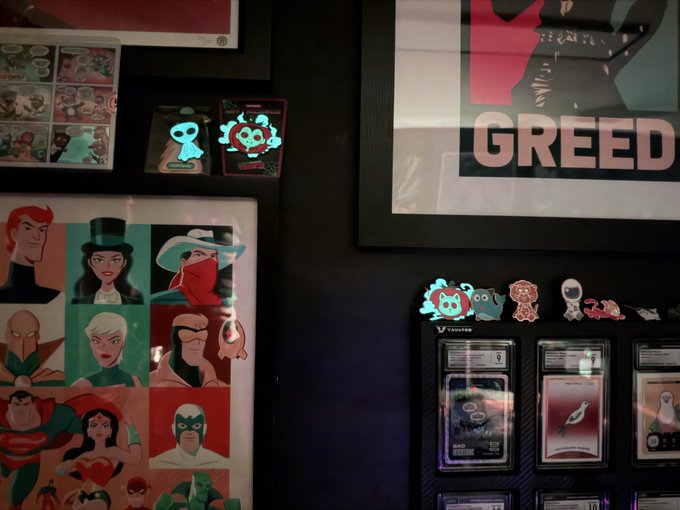 A display wall featuring framed artwork, including a Justice League character poster, a 'GREED' poster, and collectible cards or patches.