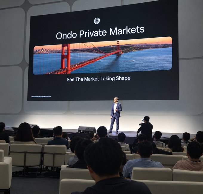 A presenter in a suit stands on stage at a tech/business conference presenting 'Ondo Private Markets - See The Market Taking Shape,' with a large screen showing the Golden Gate Bridge and an audience seated in the foreground.