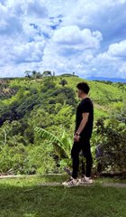 A young man in a black t-shirt and dark pants stands on a grassy ridge overlooking lush green rolling hills and a bright cloudy sky.