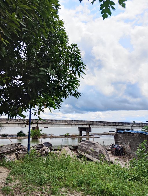 An outdoor scene featuring a concrete bridge over water, with old wooden boats resting on the shore beneath a cloudy sky and green tree foliage.