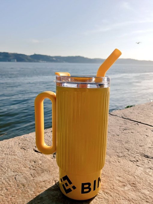 A tall yellow tumbler with a matching straw sits on a stone ledge overlooking the ocean, displaying "BIN" text on its side as part of a "Gm!" morning greeting by the sea.