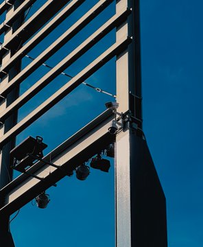 A low-angle shot looking up at the metal framework of a stadium or large structure with stage lights mounted on it against a deep blue sky.
