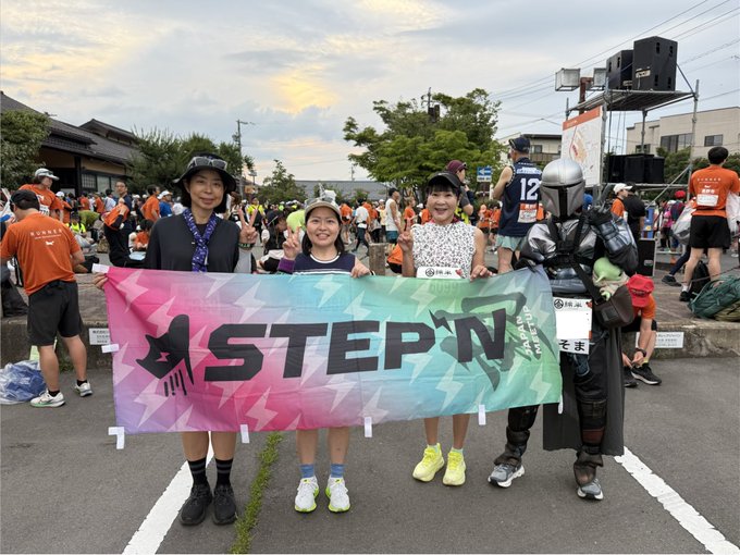 Three people pose holding a colorful 'STEP N' banner at an outdoor running event, with one person on the right dressed in a Mandalorian-style silver armor costume.