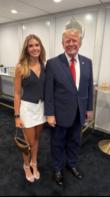 A young woman in a black vest and white mini skirt poses for a photo with Donald Trump, who is wearing his signature blue suit and red tie.